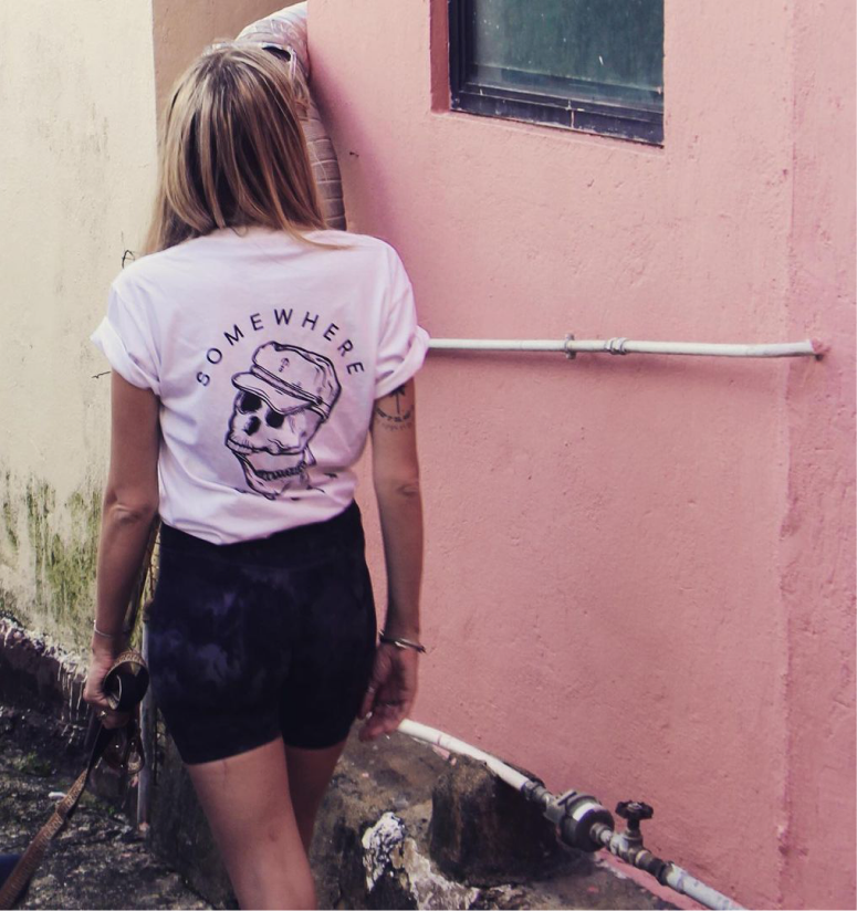 Woman walking past a pink wall wearing Somewhere South skull t-shirt
